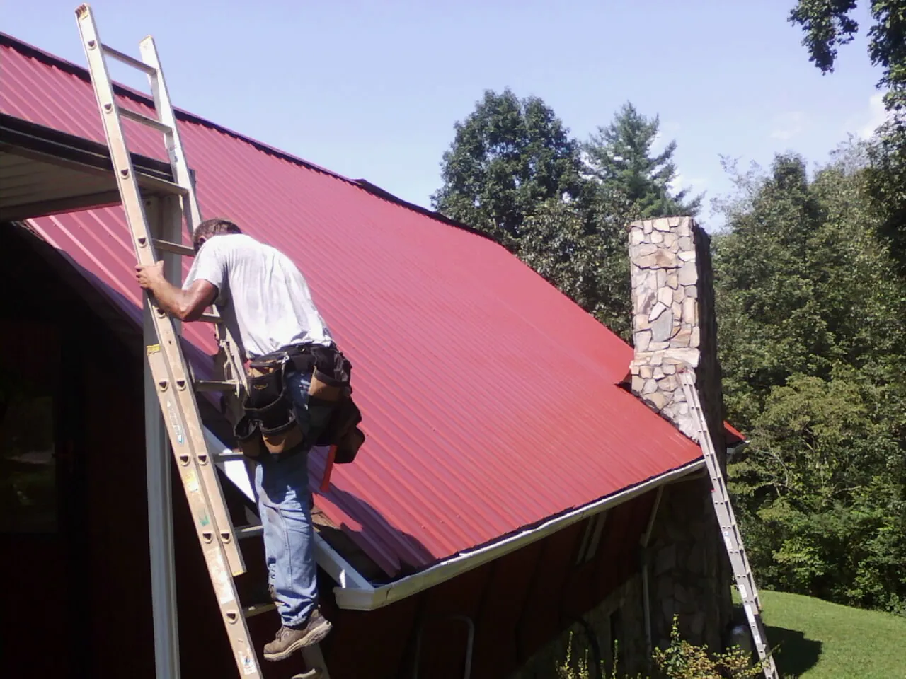Roofer on a ladder beside a red metal roof and stone chimney — Soffit & Fascia Repair by Roofing Brothers in Adamsburg